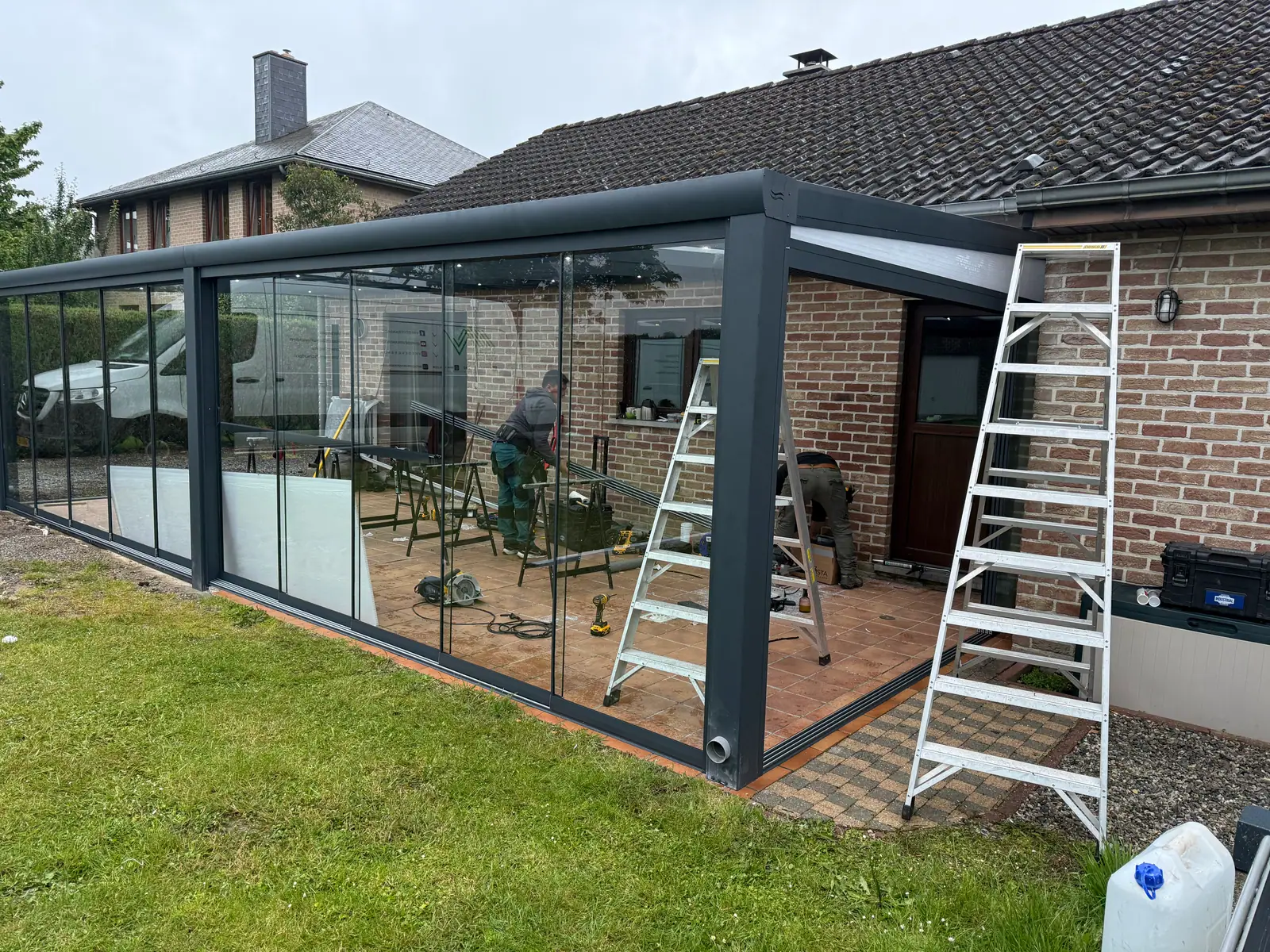 Zwei Monteure von SAVA Glass bauen bei Kunden zu Hause eine Terrassenüberdachung auf, mit Leitern und Werkzeug